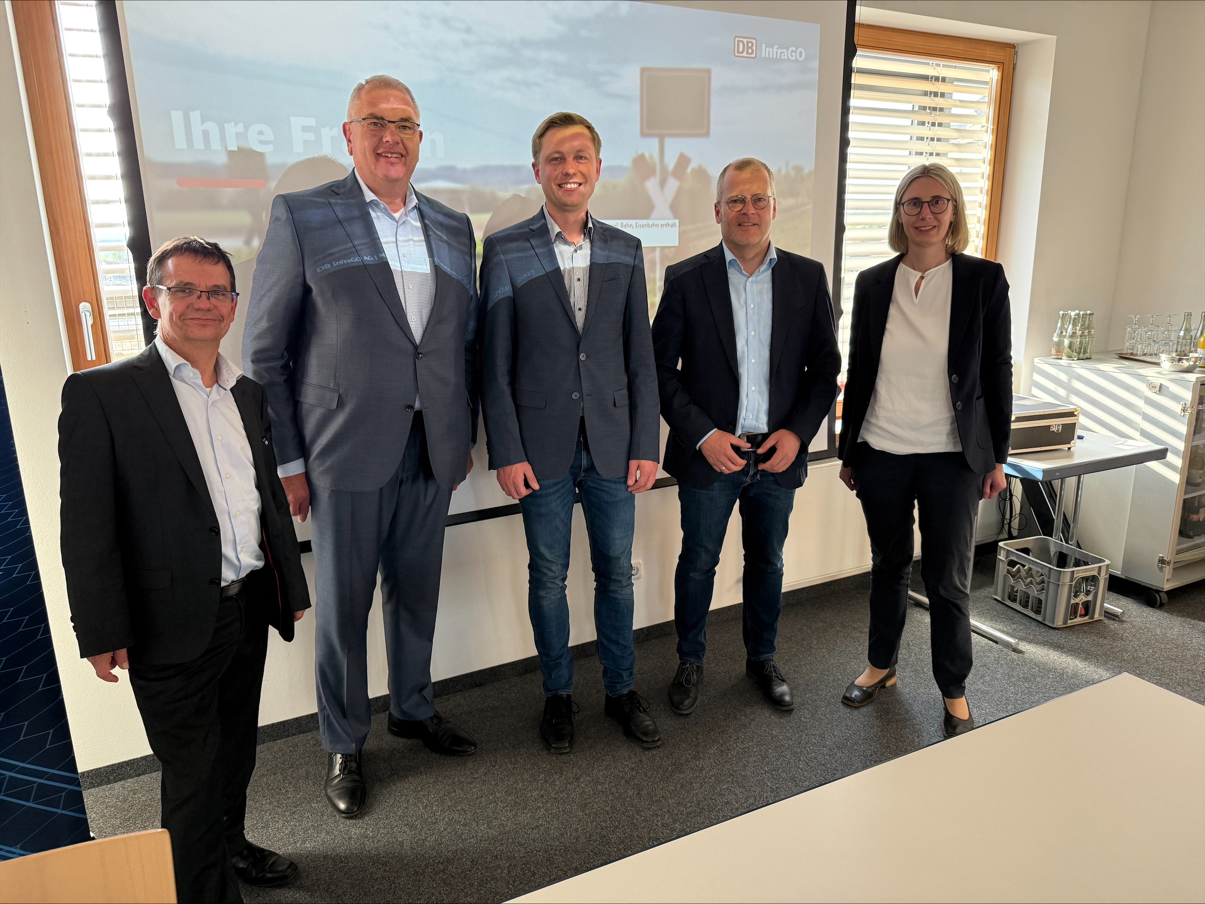 Fünf Personen stehen in einem Raum vor einer Präsentation zu einem Gruppenbild zusammen.