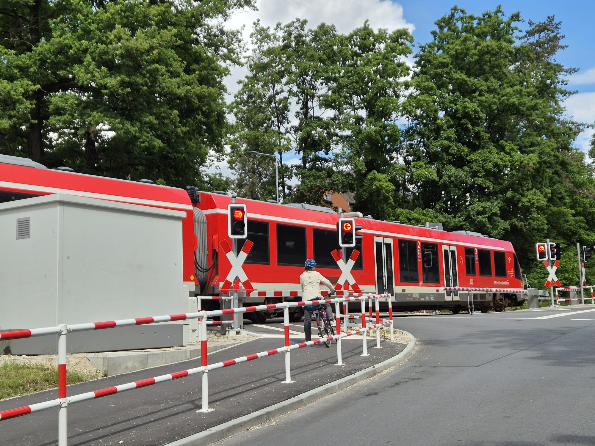 Zu sehen ist der neue Bahnübergang, ein Zug der vorbeifährt, sowie eine Person auf dem Fahrrad die am Bahnübergang wartet